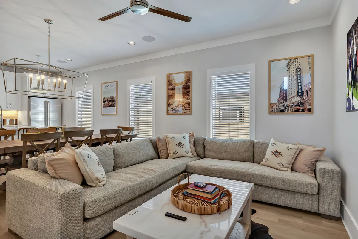 A welcoming living room with a large sectional couch, decorative artwork, a coffee table, and an open dining area visible in the background. The layout is designed for both relaxation and entertaining, with clear sightlines that reinforce the open-concept feel.