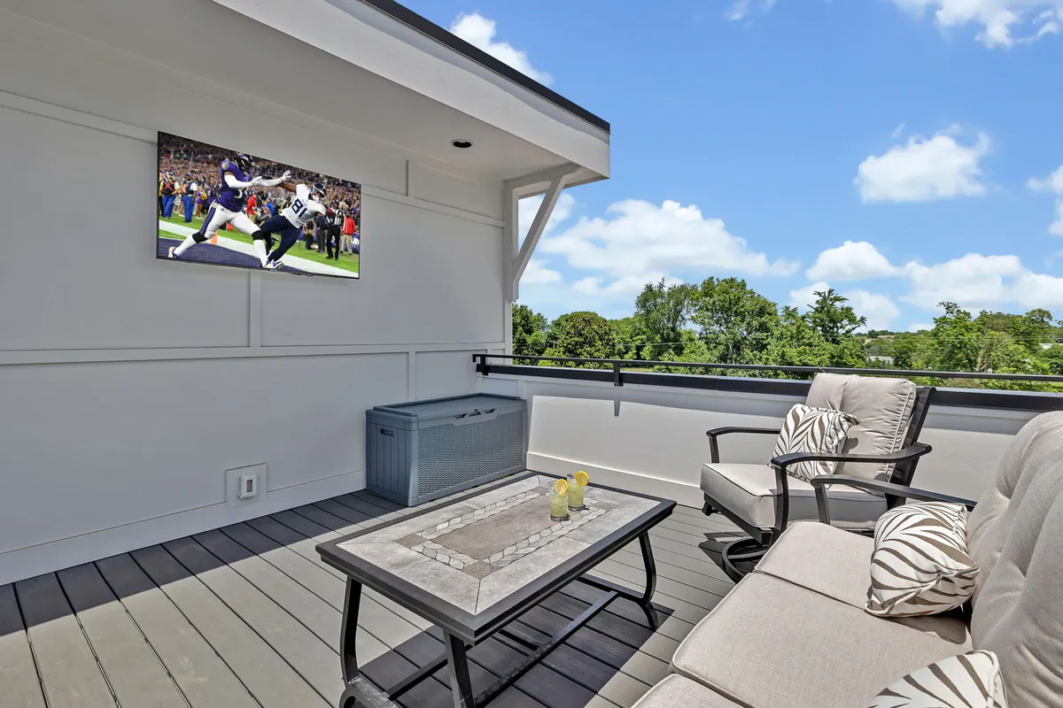 A rooftop patio featuring a stylish outdoor seating area, a television screen mounted on the exterior wall, and a lush green backdrop. The space exudes comfort and relaxation, designed for both socializing and entertainment in an open-air environment.