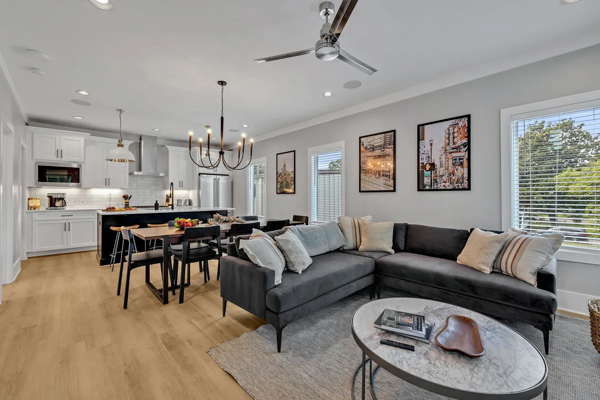 A spacious, elegantly furnished open-concept living and dining area with a plush sectional sofa, chic dining table under a sparkling chandelier, modern kitchen island, and artistic wall décor. The room is bright and inviting, perfect for group gatherings.