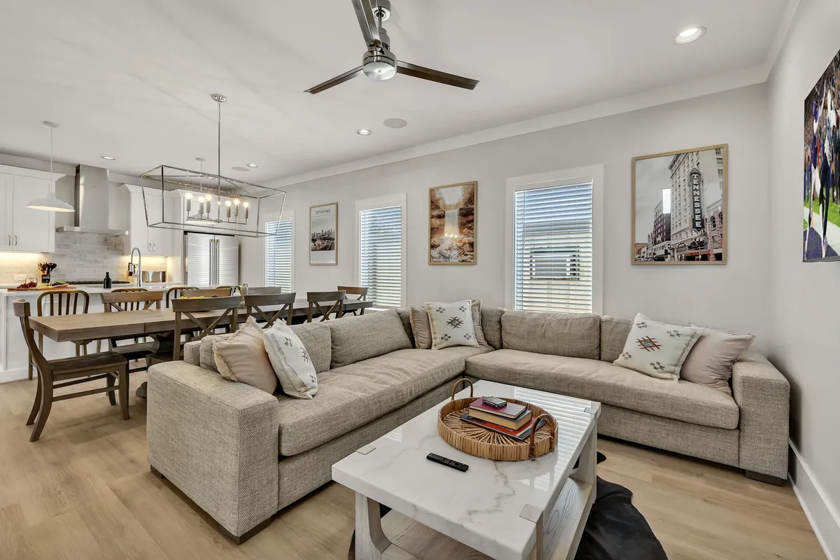 A spacious open-concept living and dining area featuring a large sectional sofa, a formal dining table set for eight, elegant chandeliers above, and tasteful artwork on the walls. The seating is arranged for both group conversations and cozy movie nights, with ample room for guests to gather.