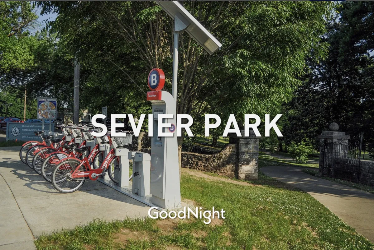 An exterior scene of a bicycle rental station near park signage, surrounded by lush greenery. The setting is active and communal, ideal for family outings, biking adventures, and social gatherings in a local park environment.